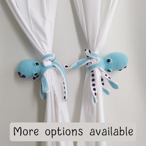 Può includere: Due fermatenda a forma di polpo all'uncinetto in azzurro con dettagli viola. I fermatenda a forma di polpo tengono indietro le tende bianche.