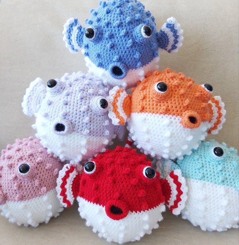Puede incluir: Seis juguetes de pez globo de crochet en varios colores, incluyendo azul, morado, naranja, rosa, rojo y turquesa. Cada juguete tiene detalles blancos y ojos negros.
