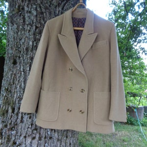 Könnte beinhalten: Ein beigefarbener, zweireihiger Blazer mit goldenen Knöpfen und zwei Vordertaschen. Die Jacke hängt an einem Holzbügel vor einem Baumstammhintergrund. Das Innenfutter hat ein dunkelblaues und braunes Muster.