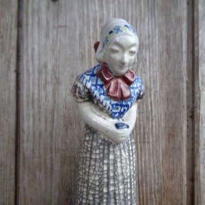 May include: A ceramic figurine of a woman wearing a blue and white patterned dress and a red bow. The figurine has a cracked glaze finish.