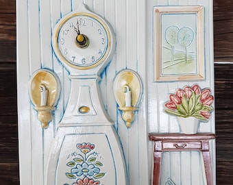May include: A white ceramic wall clock with a blue and white floral design. The clock has a white face with black Roman numeral markings and a gold-coloured hour and minute hand. The clock is surrounded by a white frame with a blue border. The clock is mounted on a white wall with a blue and white striped pattern. There is a small white table with a brown top and a basket of yarn on the table. There is a picture frame with a landscape scene on the wall. The clock is a decorative piece that would add a touch of charm to any home.