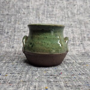 Op de afbeelding: Een kleine keramische pot met twee zijhandvatten, voorzien van een gespikkeld groen geglazuurd bovendeel en een effen donkerbruine ongeglazuurde basis. De pot heeft een wijde opening en een afgerond lichaam met contrasterende texturen.
