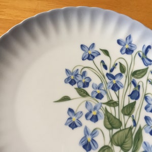 May include: A white porcelain plate with a scalloped edge and a blue gradient border. The plate is decorated with a cluster of blue violet flowers and green leaves.