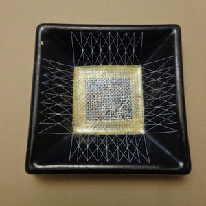 May include: A black square ceramic dish with a white geometric design and a gold square in the center.