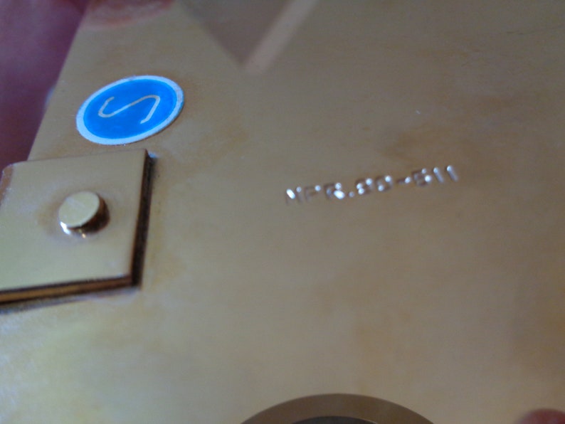K&ouml;nnte beinhalten: Nahaufnahme eines goldfarbenen Metallobjekts mit einem blauen Aufkleber, auf dem "S" steht, und einem kleinen Quadrat mit einem runden Knopf. Das Objekt tr&auml;gt die Gravur "HP-6.80-811".
