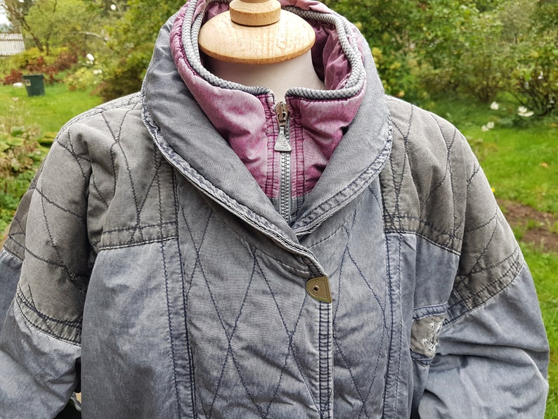 May include: A grey quilted jacket with a pink lining and a zip closure. The jacket has a diamond-shaped quilted pattern and a button on the front.
