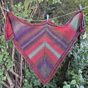 May include: A triangular knit shawl featuring a variegated color pattern of red, purple, green, and brown. The garment is constructed with a central vertical eyelet stitch detail and is suspended from a white clothesline by three blue clips.