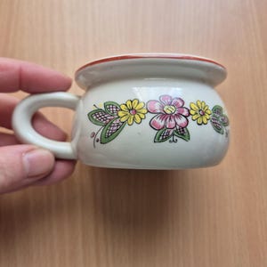 May include: A small, white ceramic cup with a handle, decorated with colourful floral designs. The rim is trimmed with a red band. The cup is likely used for drinking beverages.