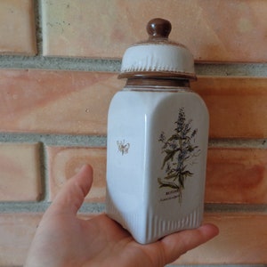 Può includere: Barattolo in ceramica bianca con coperchio marrone e design floreale. Il barattolo ha un design a farfalla da un lato e la parola "Melissa" dall'altro.