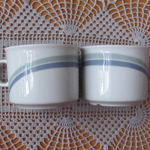 May include: Two white ceramic mugs with a blue and green stripe design. The mugs are sitting on a crocheted doily.