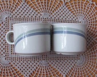 Peut inclure: Deux tasses en céramique blanche avec un motif de rayures bleues et vertes. Les tasses sont posées sur un napperon en crochet.