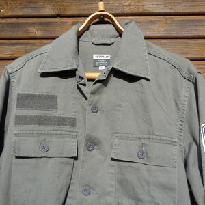 Puede incluir: Una camisa de trabajo verde utilitaria con un parche de logotipo de Caterpillar y una etiqueta m&aacute;s peque&ntilde;a que dice "Caterpillar Durable Workwear". La camisa tiene dos bolsillos grandes en la parte delantera y un bolsillo m&aacute;s peque&ntilde;o en la manga izquierda.