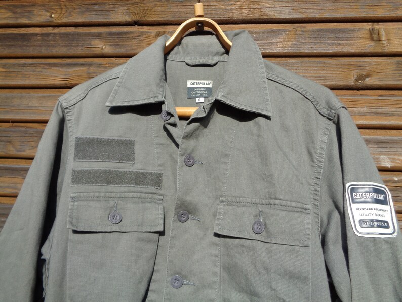 Puede incluir: Una camisa de trabajo verde utilitaria con un parche de logotipo de Caterpillar y una etiqueta m&aacute;s peque&ntilde;a que dice "Caterpillar Durable Workwear". La camisa tiene dos bolsillos grandes en la parte delantera y un bolsillo m&aacute;s peque&ntilde;o en la manga izquierda.