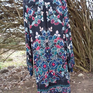 May include: A long-sleeved black dress with a vibrant floral pattern in red, blue, and teal. The dress features a button-down front and a decorative border at the hemline. The dress is displayed on a wooden mannequin.