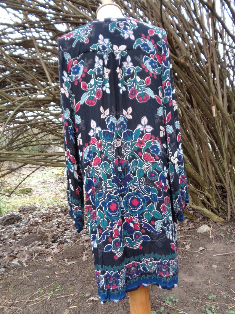 May include: A long-sleeved black dress with a vibrant floral pattern in red, blue, and teal. The dress features a button-down front and a decorative border at the hemline. The dress is displayed on a wooden mannequin.