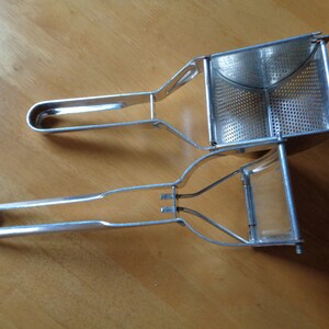 May include: Two stainless steel potato ricers on a wooden surface. The ricers have a perforated basket and long handles with rounded ends. The handles are connected to the basket with a metal frame.