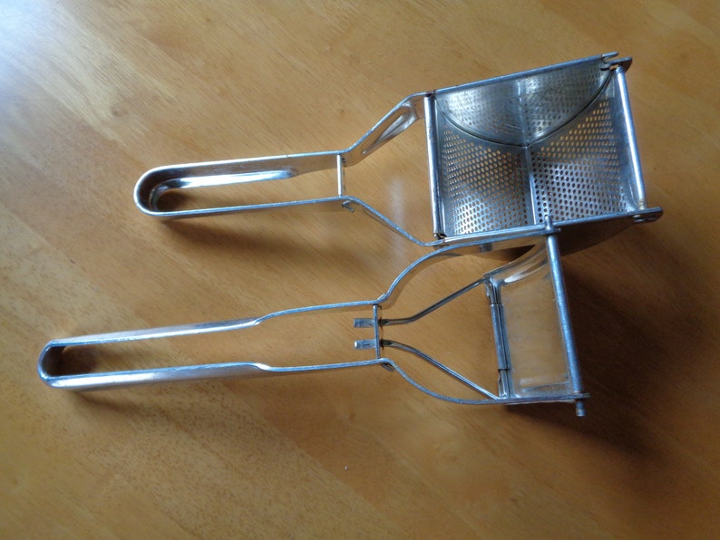 May include: Two stainless steel potato ricers on a wooden surface. The ricers have a perforated basket and long handles with rounded ends. The handles are connected to the basket with a metal frame.