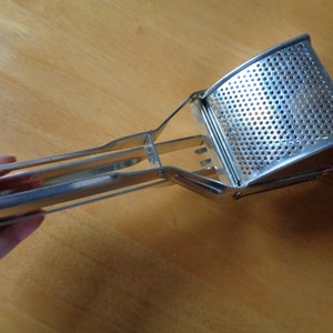 May include: A stainless steel potato masher. It has a perforated mashing head and a long handle. The masher is designed with a curved head and a handle for easy grip and use. The metal has a shiny, reflective surface.