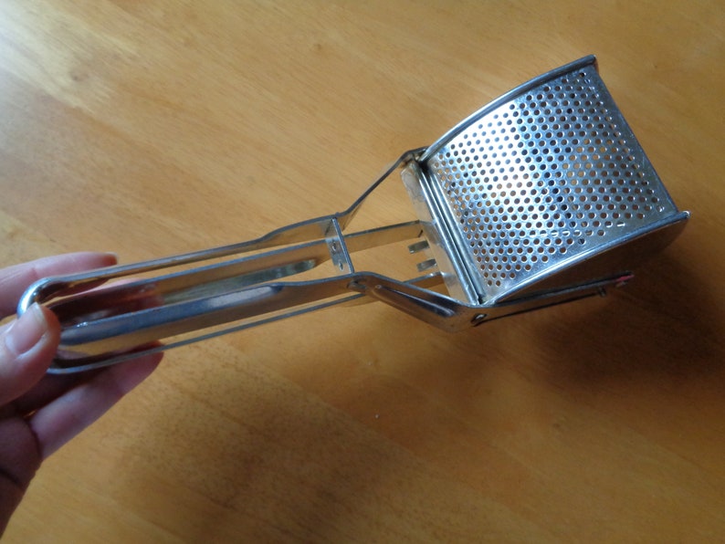 May include: A stainless steel potato masher. It has a perforated mashing head and a long handle. The masher is designed with a curved head and a handle for easy grip and use. The metal has a shiny, reflective surface.