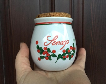 May include: White ceramic jar with a cork lid and a red and green floral design. The jar says "Senap" which is Swedish for mustard.