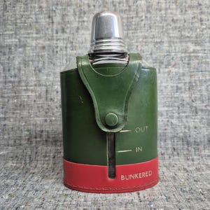 Op de afbeelding: Een vintage flacon met een zilveren dop en een groene leren hoes. De hoes heeft een rode basis en een riem met een knoopsluiting. De woorden "OUT", "IN" en "BUNKERED" zijn zichtbaar op de flacon.