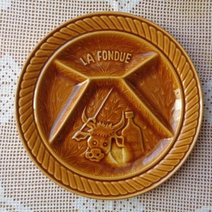May include: A brown ceramic fondue dish with four sections. The dish has a rope-like border and a bull illustration in the center. The text "LA FONDUE" is written in the top section.