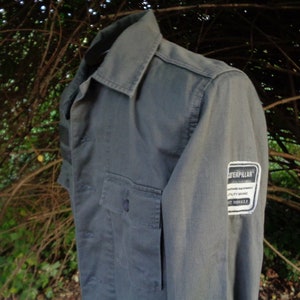 Puede incluir: Una camisa de trabajo gris de manga larga con un parche de la marca Caterpillar en la manga. El parche es blanco con texto negro.