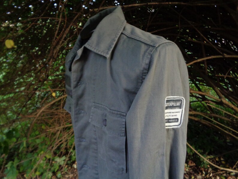 Puede incluir: Una camisa de trabajo gris de manga larga con un parche de la marca Caterpillar en la manga. El parche es blanco con texto negro.