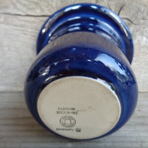 May include: A blue ceramic mug with a white bottom. The bottom of the mug has the text "STINCOOS SWEDEN" and a logo.