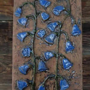 K&ouml;nnte beinhalten: Eine Keramik-Wandplatte mit einem Reliefdesign aus blauen Glockenblumen auf braunem Hintergrund.