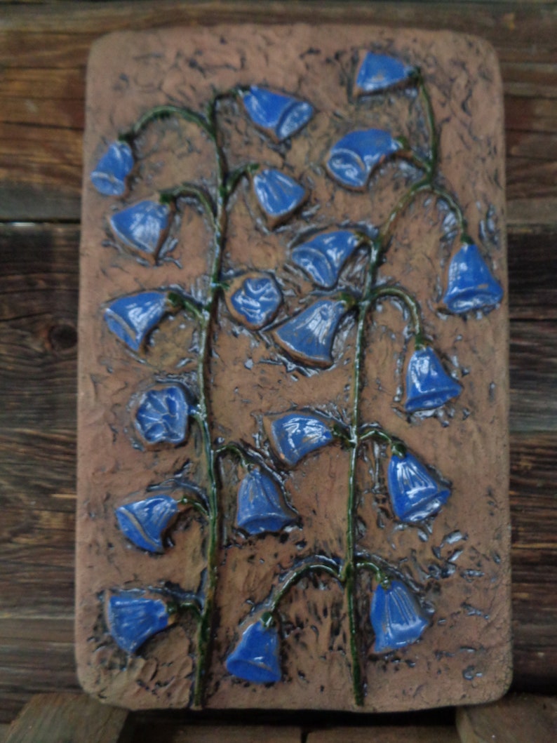 K&ouml;nnte beinhalten: Eine Keramik-Wandplatte mit einem Reliefdesign aus blauen Glockenblumen auf braunem Hintergrund.