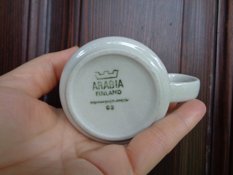 Puede incluir: Una peque&ntilde;a taza de cer&aacute;mica blanquecina con un asa redondeada. La base de la taza muestra el texto "ARABIA FINLAND" y "DISHWASHER-PROOF 63" en verde. La taza est&aacute; sujeta en una mano.