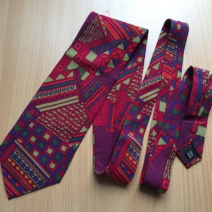 May include: A red, green, purple and orange patterned tie with a geometric design. The tie is made of silk and has a woven label that says "100% Silk".