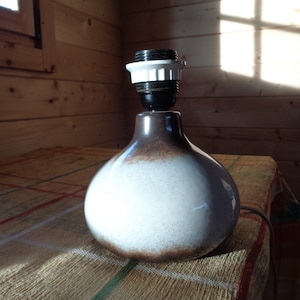 May include: A ceramic table lamp with a bulb socket. The lamp has a rounded, bulbous base with a gradient color scheme, transitioning from brown to white. The lamp sits on a patterned tablecloth in a room with wooden walls.