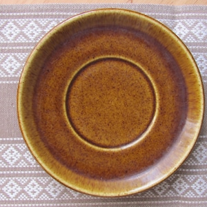 Peut inclure: Une soucoupe en céramique brune avec un centre légèrement surélevé. La soucoupe a un aspect rustique et moucheté.