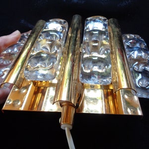 K&ouml;nnte beinhalten: Eine goldfarbene Wandleuchte mit klaren Glaspaneelen. Die Leuchte hat ein modernes Design und ist perfekt, um jedem Raum einen Hauch von Eleganz zu verleihen.