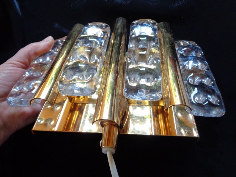 K&ouml;nnte beinhalten: Eine goldfarbene Wandleuchte mit klaren Glaspaneelen. Die Leuchte hat ein modernes Design und ist perfekt, um jedem Raum einen Hauch von Eleganz zu verleihen.