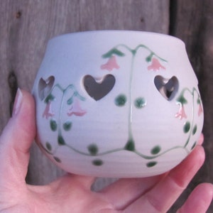 May include: A white ceramic candle holder with heart-shaped cutouts and floral accents. The holder features pink bell-shaped flowers and green vines. The candle holder is held in a hand.