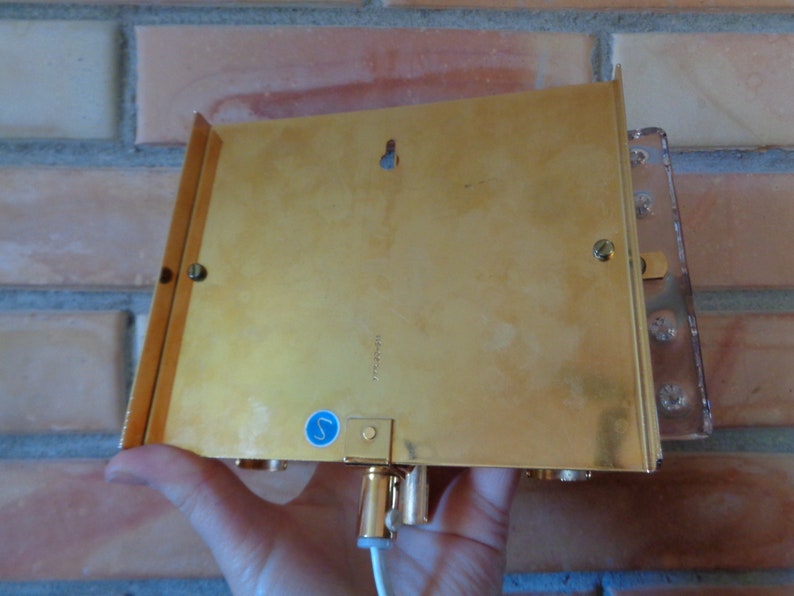 K&ouml;nnte beinhalten: R&uuml;ckansicht einer goldfarbenen Wandleuchte mit einem klaren Glasschirm. Die Leuchte hat einen kleinen blauen Aufkleber auf der R&uuml;ckseite mit dem Buchstaben "S" darauf.