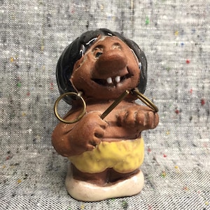 May include: A ceramic figurine of a smiling person with brown skin, black hair, and yellow shorts. The figurine is wearing large hoop earrings and holding a stick in its hand.