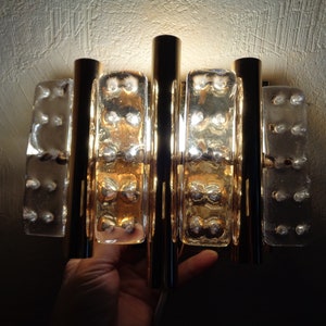 K&ouml;nnte beinhalten: Eine Wandleuchte aus Gold und klarem Glas mit geometrischem Design. Die Leuchte hat vier vertikale St&auml;be und drei rechteckige Glasscheiben. Die Glasscheiben haben eine strukturierte Oberfl&auml;che und werden von hinten beleuchtet.