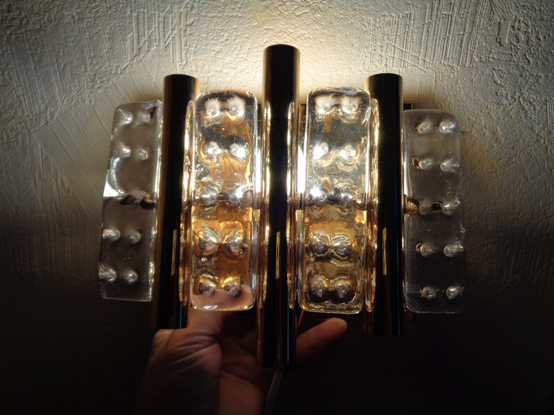 K&ouml;nnte beinhalten: Eine Wandleuchte aus Gold und klarem Glas mit geometrischem Design. Die Leuchte hat vier vertikale St&auml;be und drei rechteckige Glasscheiben. Die Glasscheiben haben eine strukturierte Oberfl&auml;che und werden von hinten beleuchtet.