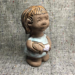 May include: A ceramic figurine of a girl with brown hair wearing a light blue dress and holding a small flower. The figurine has a rustic, hand-crafted look.