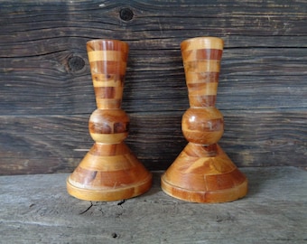 May include: Two wooden candle holders with a natural wood finish. The holders are made of multiple pieces of wood that are stacked and glued together. The holders have a round base and a cylindrical top.