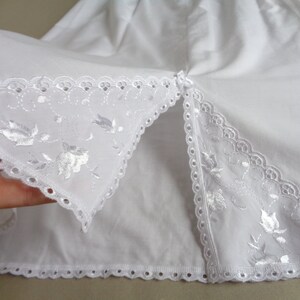 May include: Close-up of white fabric with delicate floral embroidery and scalloped edges. The fabric is folded, revealing a split opening. The embroidery features detailed floral and leaf patterns, adding a touch of elegance.