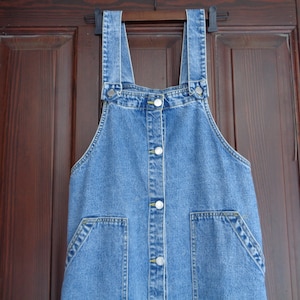 Op de afbeelding: Een blauwe denim overalljurk met een knoopsluiting aan de voorkant en twee zakken. De jurk hangt aan een houten deur.
