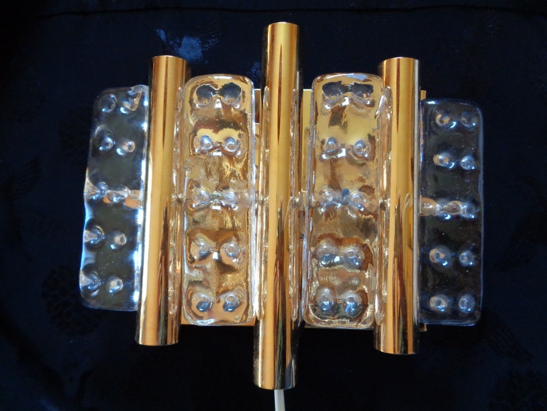 K&ouml;nnte beinhalten: Goldene Wandleuchte mit vier klaren Glasplatten, jede mit einem strukturierten Muster. Die Leuchte hat ein modernes Design und w&auml;re eine gro&szlig;artige Erg&auml;nzung f&uuml;r jedes Zuhause.