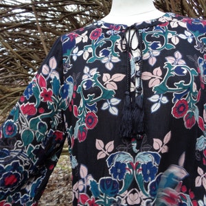May include: A black long-sleeve tunic with a floral pattern in red, blue, green, and pink. The tunic has a tie-neck detail with tassels. The sleeves are long and loose, and the fabric appears to be lightweight.