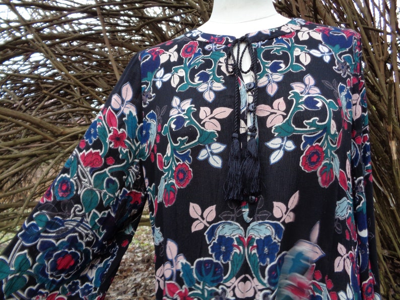 May include: A black long-sleeve tunic with a floral pattern in red, blue, green, and pink. The tunic has a tie-neck detail with tassels. The sleeves are long and loose, and the fabric appears to be lightweight.