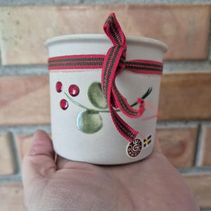 Op de afbeelding: Een kleine, crèmekleurige keramische pot met een rood en groen lint eromheen. De pot is versierd met rode bessen en groene bladeren. Een klein label met een logo en een Zweedse vlag is bevestigd.